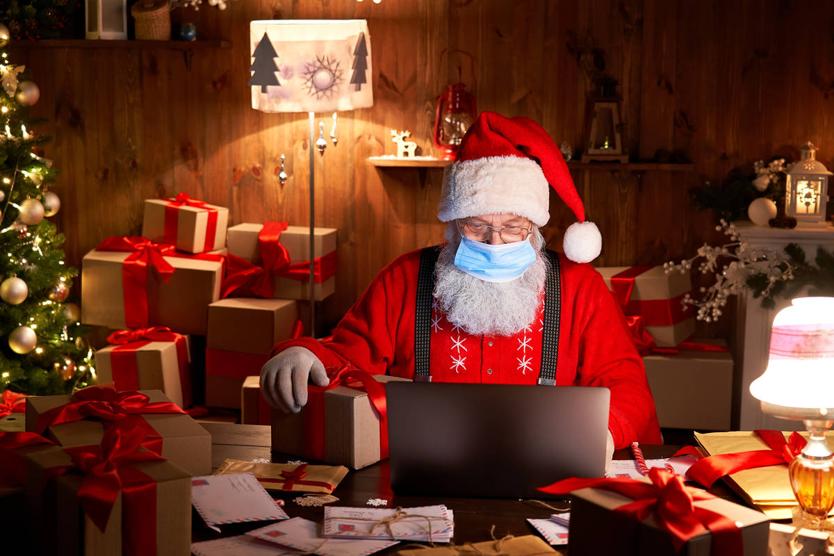 Old bearded Santa Claus wearing face mask, holding gift box on xmas eve sitting at cozy home ta ...